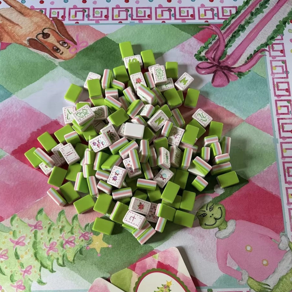Christmas Grinch mahjong tiles scattered on matching Grinch holiday game mat, with festive printed coasters, pink and green Christmas themed table setup
