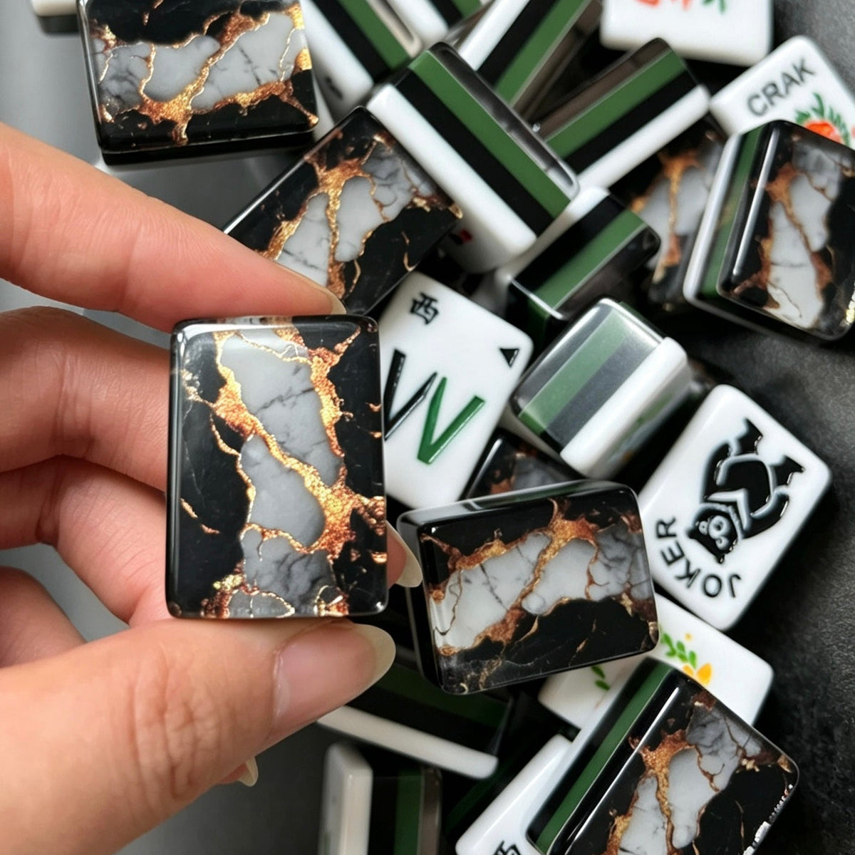 Hand holding mahjong tile displaying black marble back with white stone veins and shimmer gold foil, pile of black gold marble mahjong tiles in background