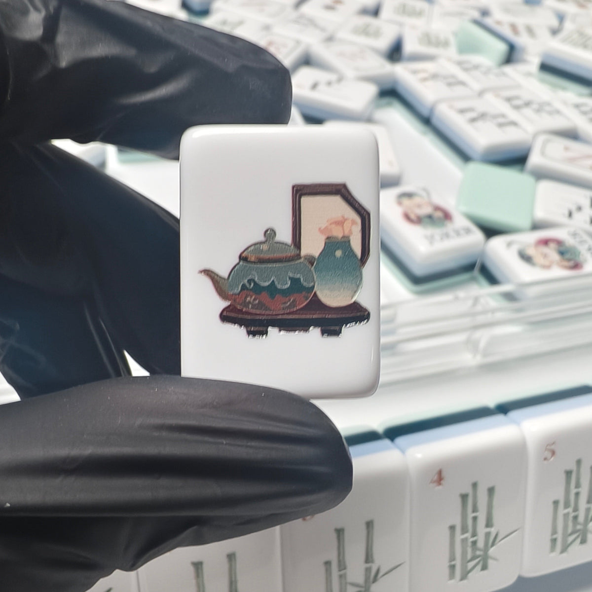 Overhead shot of scattered Chinese Sage mahjong tiles with clear acrylic tile rack on plain white background, showing mint green backs, navy-white layered edges, bamboo and house artwork on white tile fronts