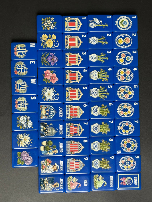 Top-down full layout of cobalt blue chinoiserie mahjong tiles arranged on black and white surface, showcasing porcelain vase bamboo, floral blooms, swan dragons and whimsical vintage joker artwork