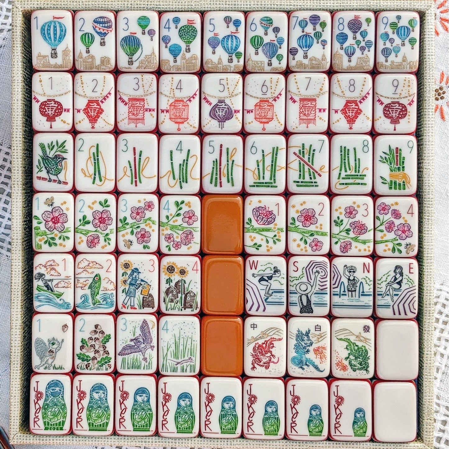Rainbow Voyage Mahjong Tiles Set — Amber‑orange backs, multi‑layer rainbow striped sides, ivory fronts with hot‑air balloon, dragon, lantern, cherry‑blossom bee and matryoshka nesting‑doll joker American mahjong tiles