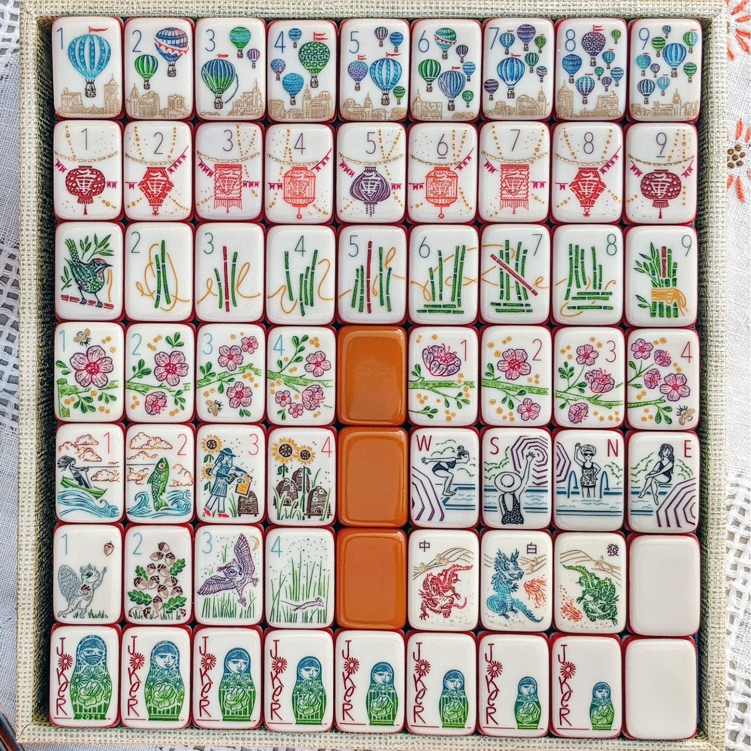 Rainbow Voyage Mahjong Tiles Set — Amber‑orange backs, multi‑layer rainbow striped sides, ivory fronts with hot‑air balloon, dragon, lantern, cherry‑blossom bee and matryoshka nesting‑doll joker American mahjong tiles