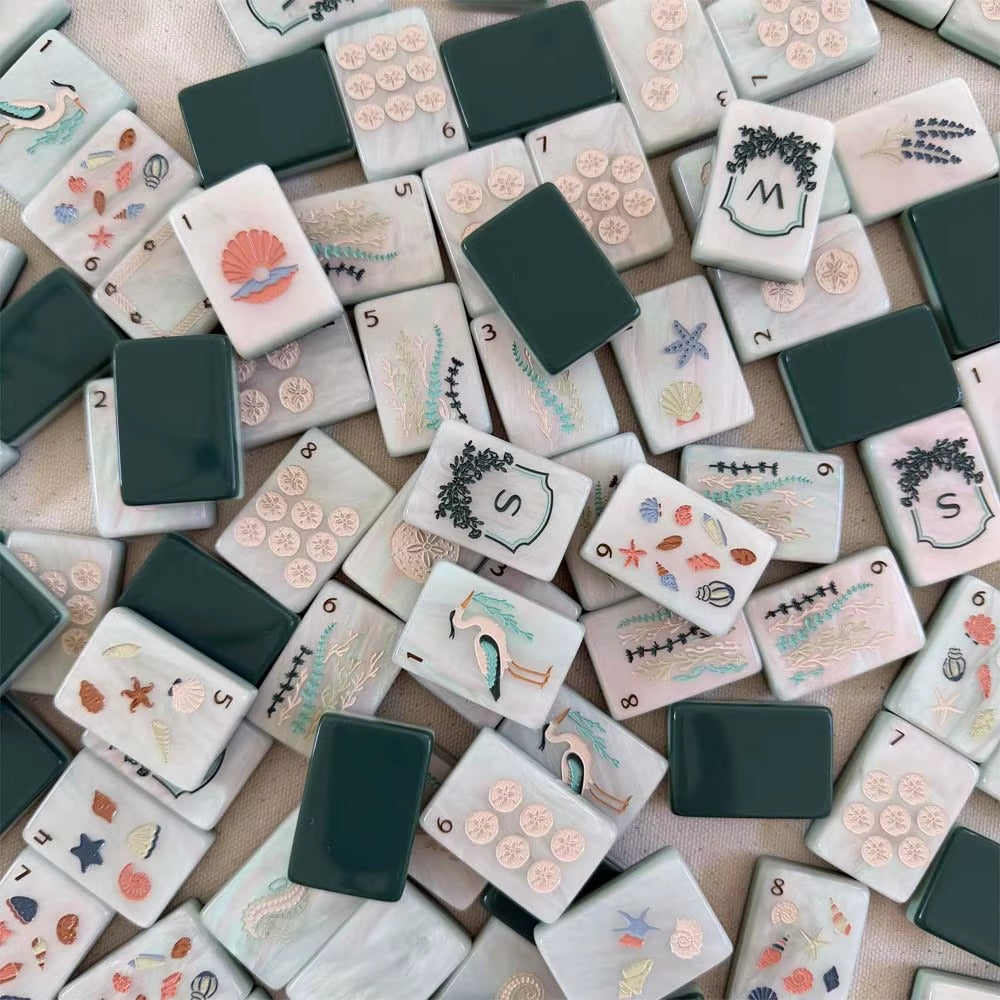 Full layout of coastal ocean American mahjong set, cream marbled tile faces with sand dollar dots, sea grass bamboo suits, seashell character tiles, nautical joker tiles and framed N E W S wind shields, deep green blank tile at corner