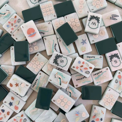 Full layout of coastal ocean American mahjong set, cream marbled tile faces with sand dollar dots, sea grass bamboo suits, seashell character tiles, nautical joker tiles and framed N E W S wind shields, deep green blank tile at corner