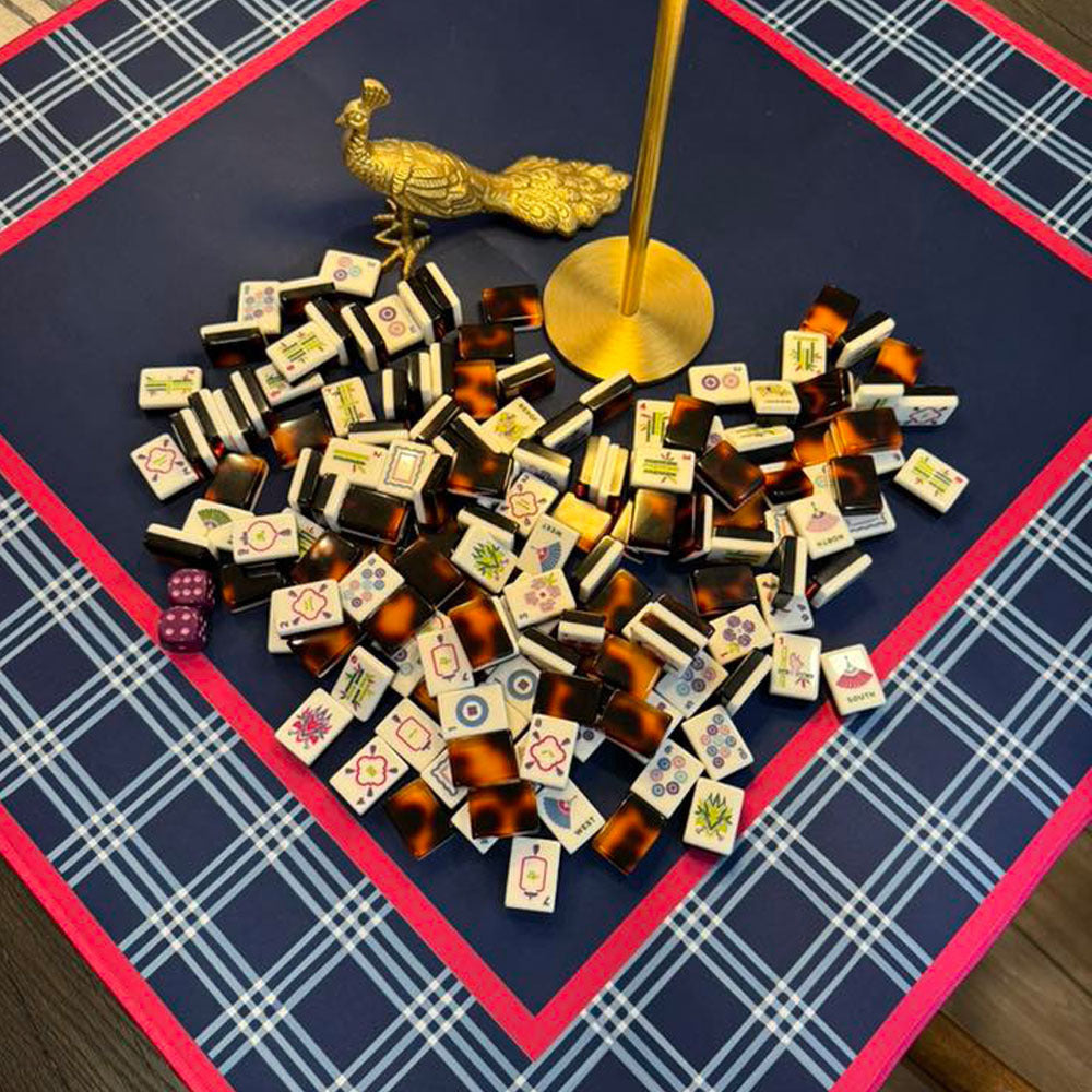Top-down view of tortoise shell American mahjong tiles scattered on navy blue plaid game mat, paired with golden peacock decor and brass lamp base, cozy game night styling