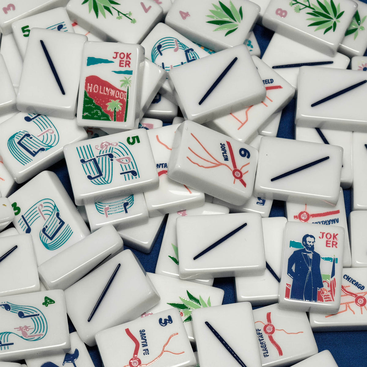 Close-up shot of American Road Trip mahjong tiles scattered on blue fabric, white resin tiles featuring Hollywood, Lincoln, music note and US highway map illustrations
