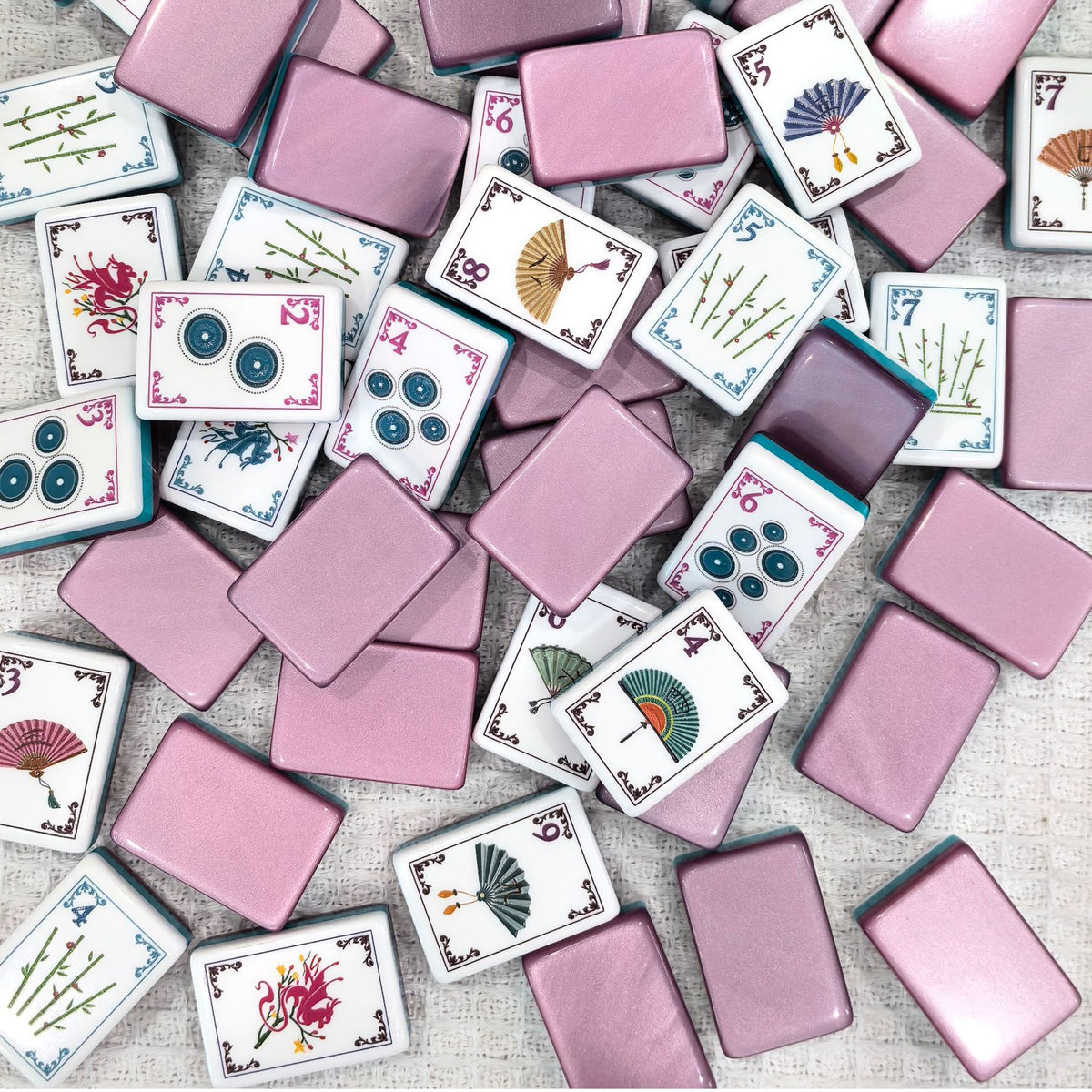 Pile of American mahjong tiles, shimmery pink backs, white tile fronts with elegant fan, bamboo, dragon and circle suit artwork, scattered on textured linen fabric