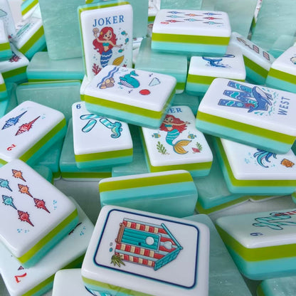 Flat lay of Mermaid Ocean American mahjong tiles, white tile faces with mermaid jokers, puffer fish dots, conch shell bamboos and nautical cracks, translucent mint green shimmer backs with aqua-lime layered stripes