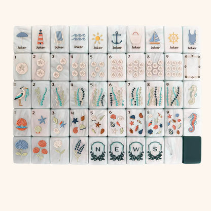 Full layout of coastal ocean American mahjong set, cream marbled tile faces with sand dollar dots, sea grass bamboo suits, seashell character tiles, nautical joker tiles and framed N E W S wind shields, deep green blank tile at corner