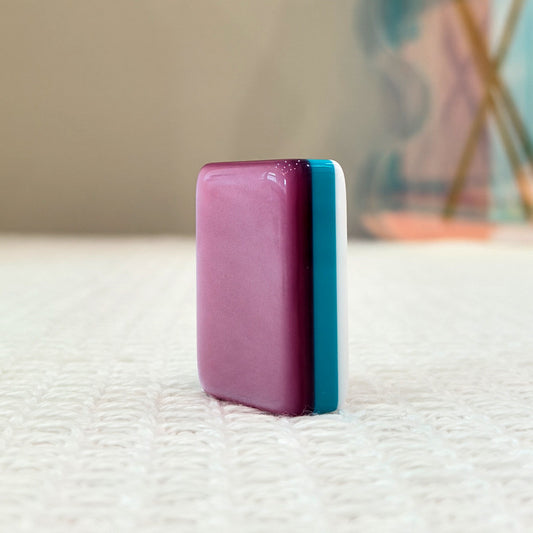 Single mahjong tile side profile, glossy shimmery pink back with teal and white layered edge, standing upright on soft white fabric