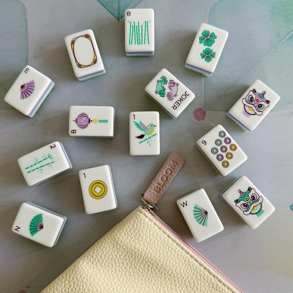 Bloom pastel mahjong tiles laid out on light blue game mat with cream leather zipper pouch tagged BLOOM, showing koi joker, lion dance, fan wind and lantern tiles