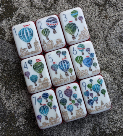 Rainbow Voyage Mahjong Tiles Set — Amber‑orange backs, multi‑layer rainbow striped sides, ivory fronts with hot‑air balloon, dragon, lantern, cherry‑blossom bee and matryoshka nesting‑doll joker American mahjong tiles