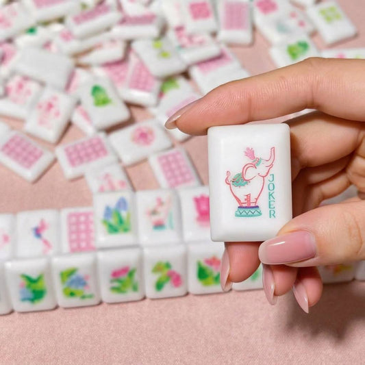 Tropical Paradise mahjong close‑up, hand holding custom elephant joker tile with pink‑green elephant graphic, blurred tropical mahjong tiles background