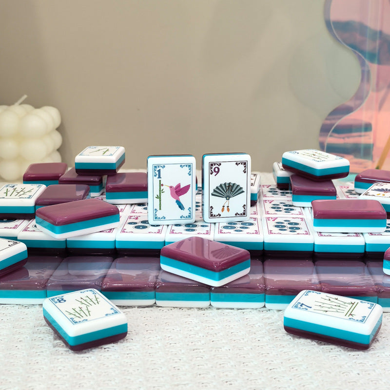 Flat lay of pink teal layered American mahjong tiles, featuring hummingbird and vintage fan illustrated tile faces, styled tabletop product shot