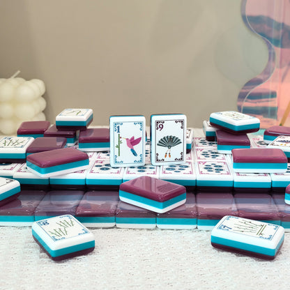 Flat lay of pink teal layered American mahjong tiles, featuring hummingbird and vintage fan illustrated tile faces, styled tabletop product shot