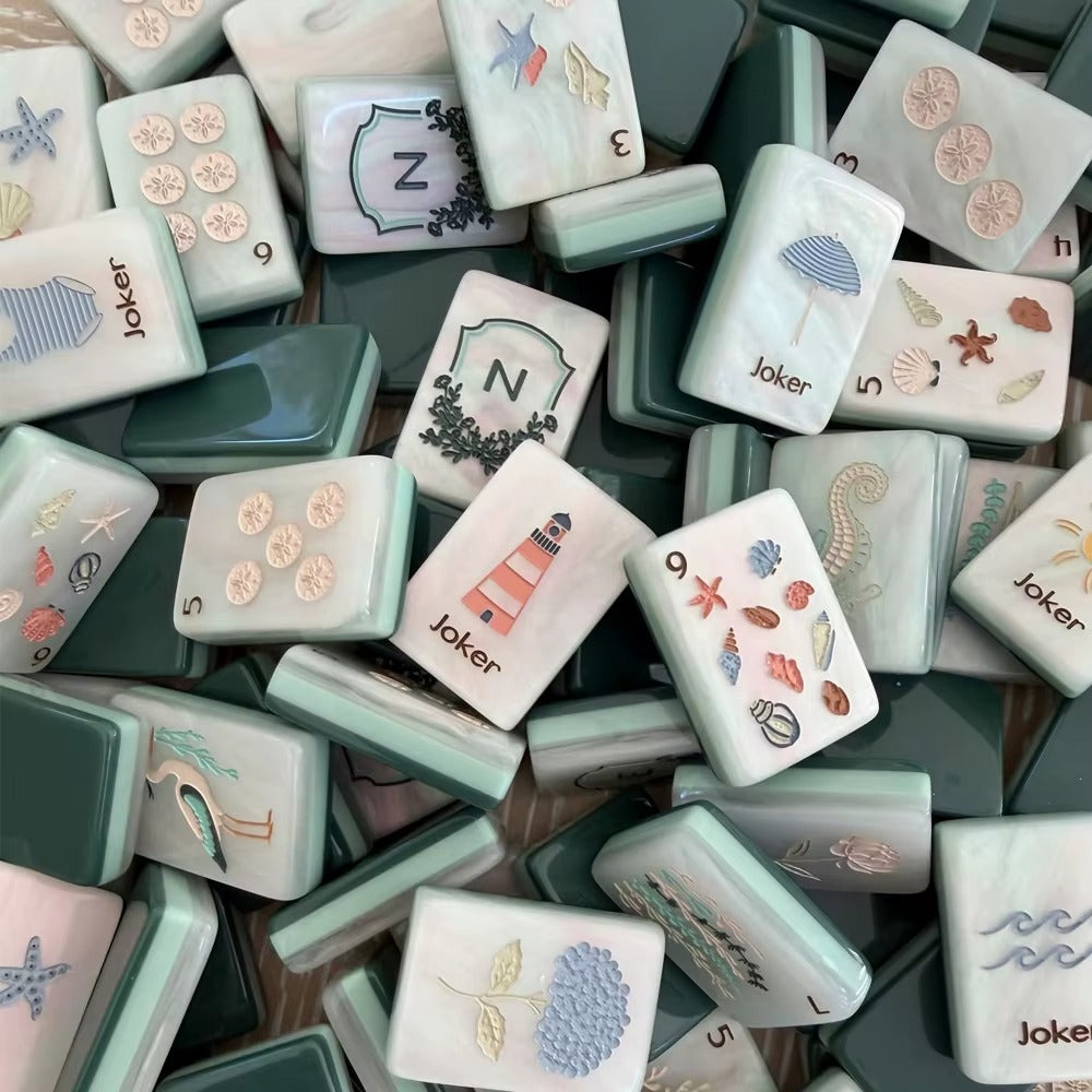 Full layout of coastal ocean American mahjong set, cream marbled tile faces with sand dollar dots, sea grass bamboo suits, seashell character tiles, nautical joker tiles and framed N E W S wind shields, deep green blank tile at corner