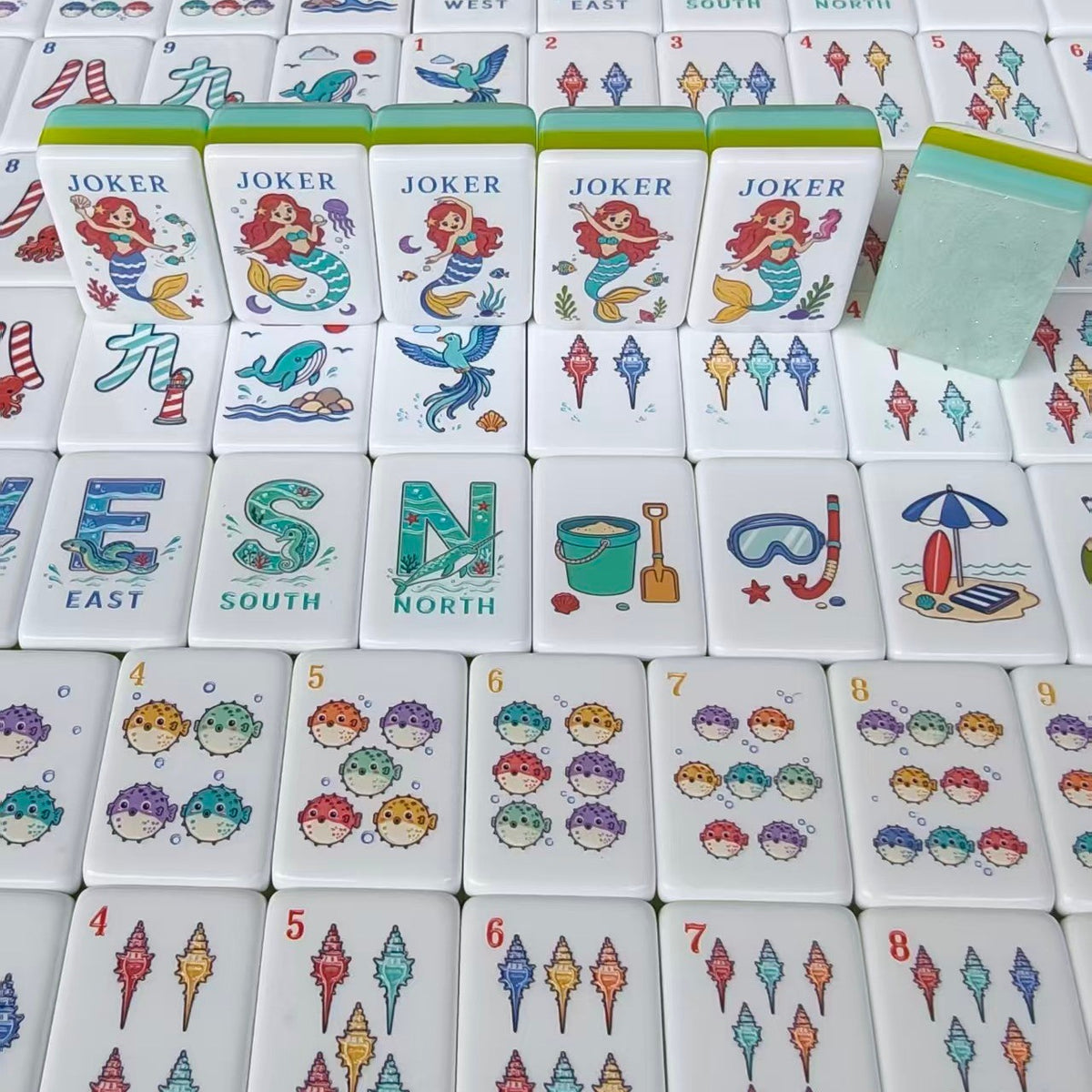 Overhead scattered shot of Mermaid Ocean mahjong tiles, showing South wind tile with seahorse, West wind with whale, North wind, ship wheel crack tiles, seahorse crack tiles, mermaid jokers and beach hut tile