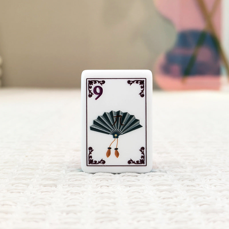 Close-up of mahjong tile front, white base with dark vintage fan graphic, number 9, decorative border, on neutral textured background