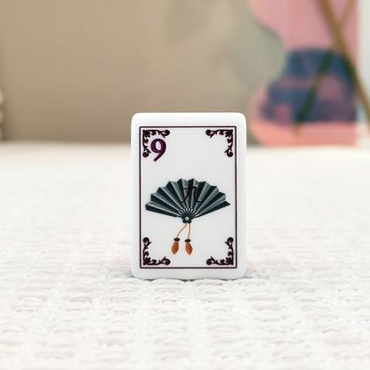 Close-up of mahjong tile front, white base with dark vintage fan graphic, number 9, decorative border, on neutral textured background