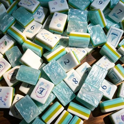 Flat top-down view of fully arranged Ocean Tide American mahjong tiles, custom nautical artwork including seashells, seaweed, sailboat and sea creature jokers, turquoise pearlescent tile backs with green-blue layered side stripes on clean white background
