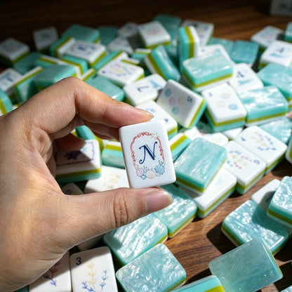 Close-up shot of hand holding North wind mahjong tile with seashell framed artwork, background filled with scattered Ocean Tide mahjong tiles with aqua pearlescent backs