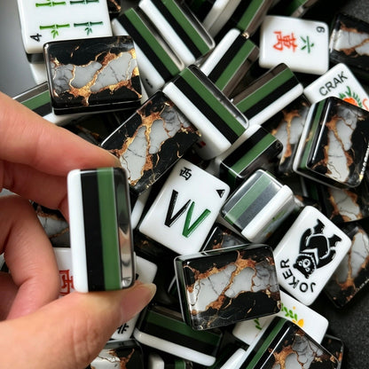 Hand holding mahjong tile displaying black marble back with white stone veins and shimmer gold foil, pile of black gold marble mahjong tiles in background