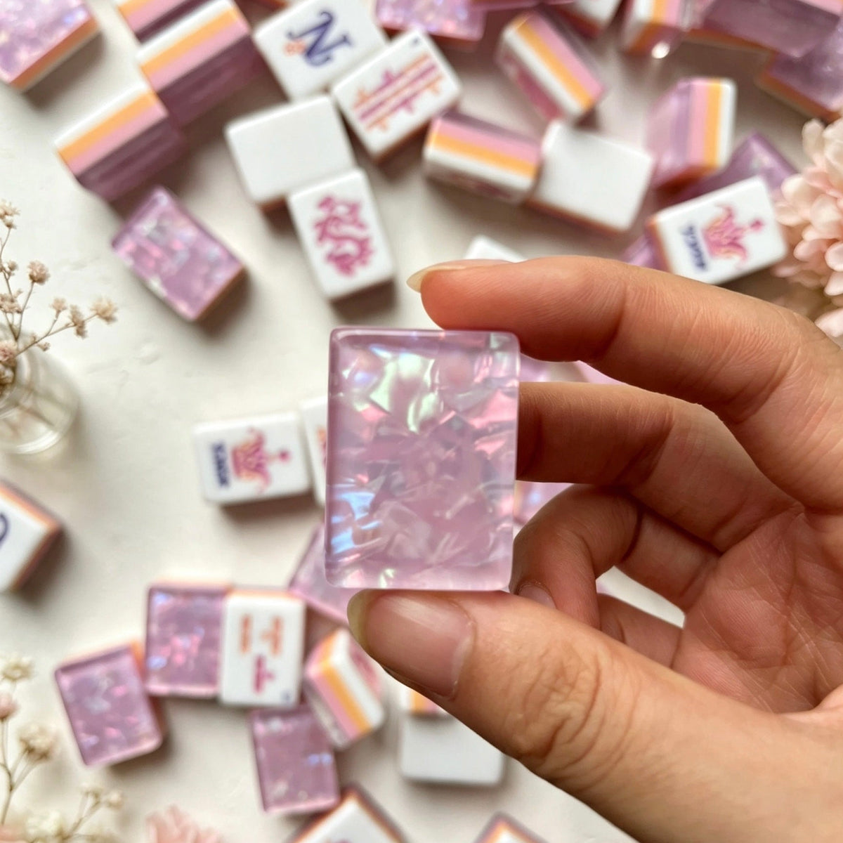 Hand holding single purple pearl resin mahjong tile, scattered purple crown mahjong set in background, floral and crown joker designs, soft floral decor