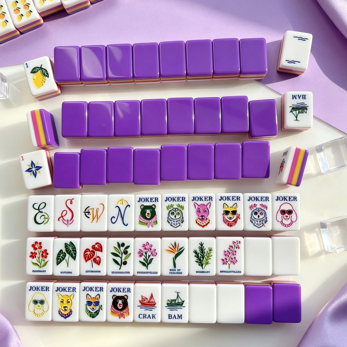 Full neatly arranged display of Purple Rainbow Striped Fruit & Fox Joker mahjong set, complete fruit, floral and animal joker tiles, styled on light stone countertop against soft purple backdrop