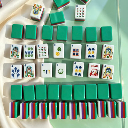 Close-up hand shot holding tropical resort mahjong 2 tile, colorful resort arch illustration, deep green backed mahjong tiles scattered in background