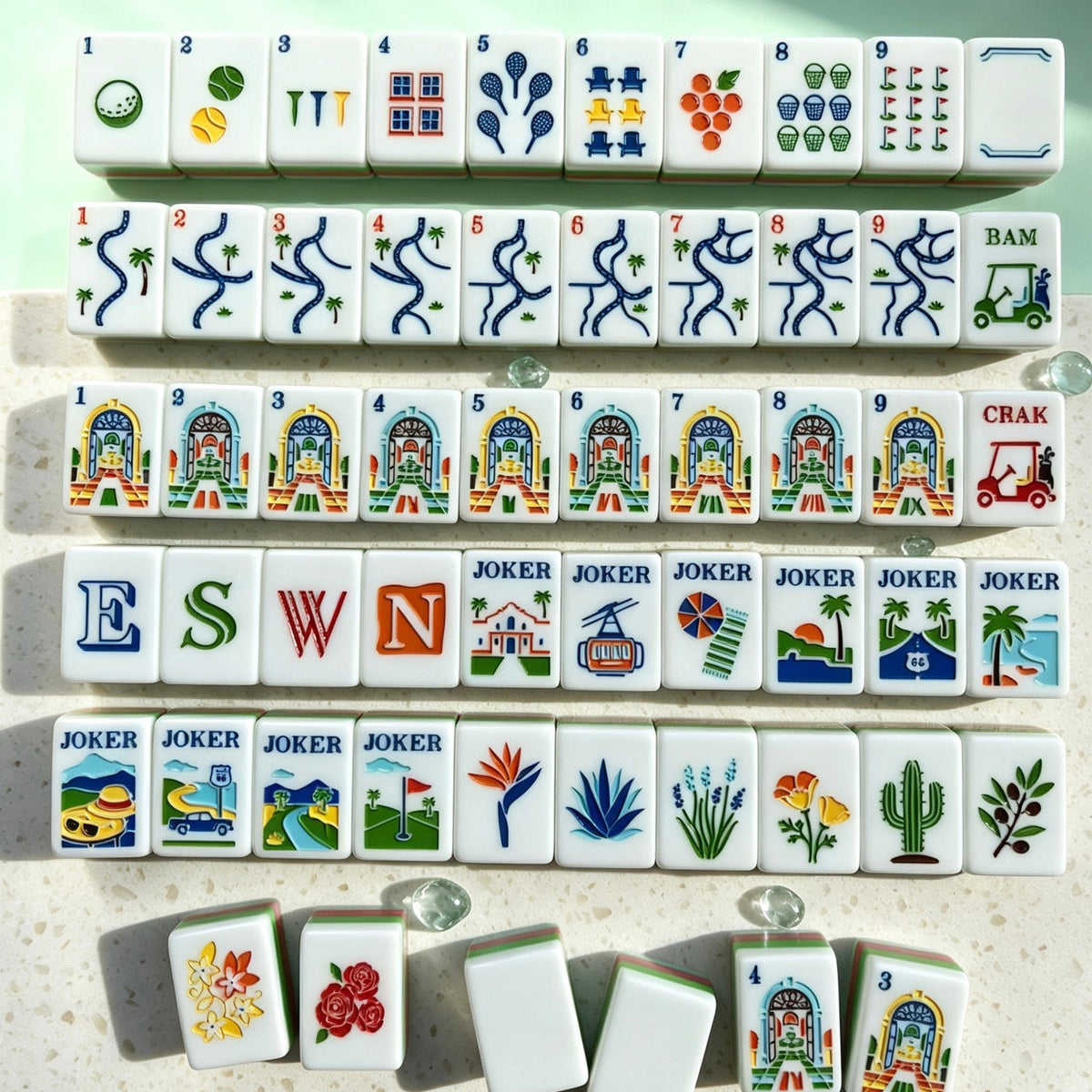 Neat full layout of Tropical Golf Resort American mahjong tiles, mint green tile backs, pink-green striped sides, custom golf, palm tree and resort building illustrations, arranged on light speckled countertop