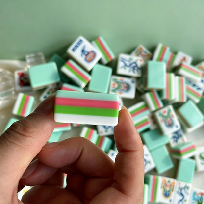 Overhead shot of scattered tropical golf resort mahjong tiles, mint green backs, pink and green striped edges, vacation themed artwork on white tile faces, styled on light green background