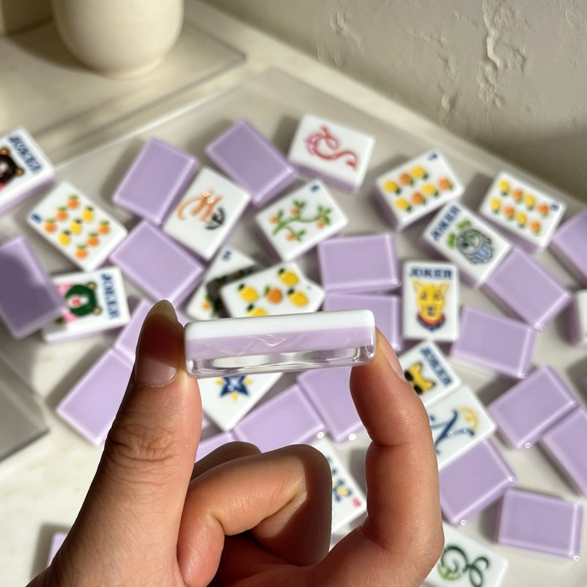 Closeup hand shot holding single layered lavender resin mahjong tile, scattered fruit floral mahjong tiles in background, cute illustrated jokers and fruit artwork