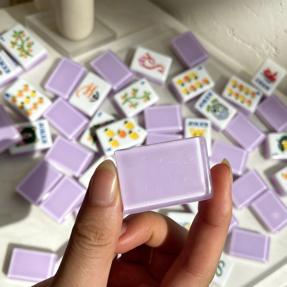 Closeup hand shot holding single layered lavender resin mahjong tile, scattered fruit floral mahjong tiles in background, cute illustrated jokers and fruit artwork