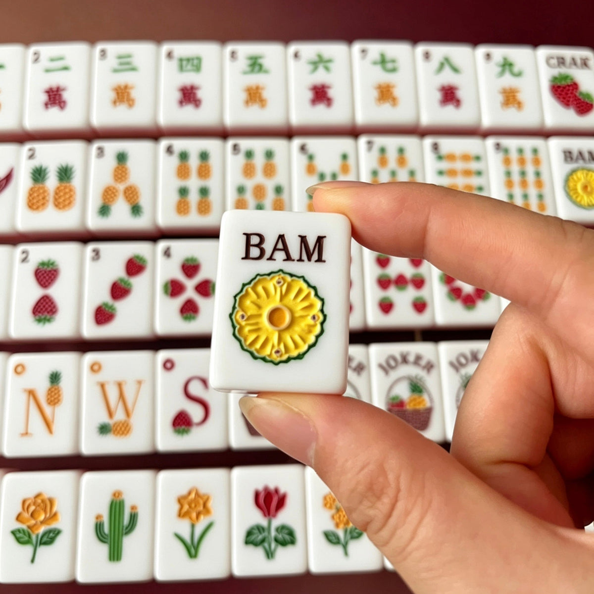 Hand holding one pink shell-back mahjong tile in front of neatly arranged tropical fruit garden mahjong set on black background, showcasing strawberry, pineapple, cactus and rose artwork