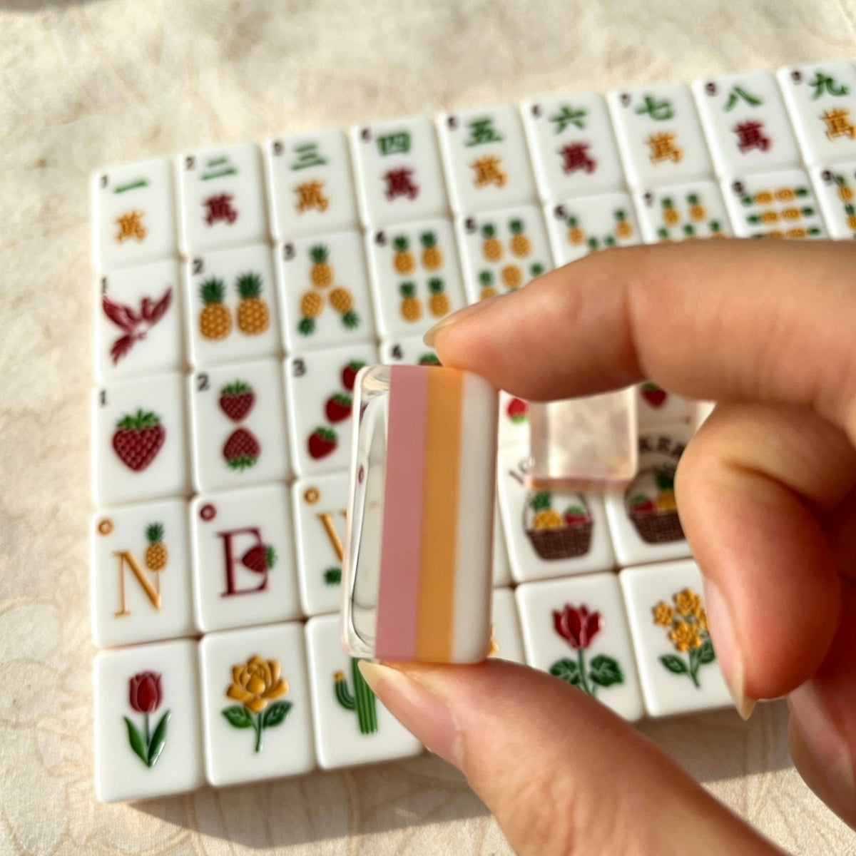Scattered pile of Fruit Garden mahjong tiles, shimmering pink pearlescent backs, pastel pink-orange layered edges, strawberry and pineapple illustrated tile faces on linen surface