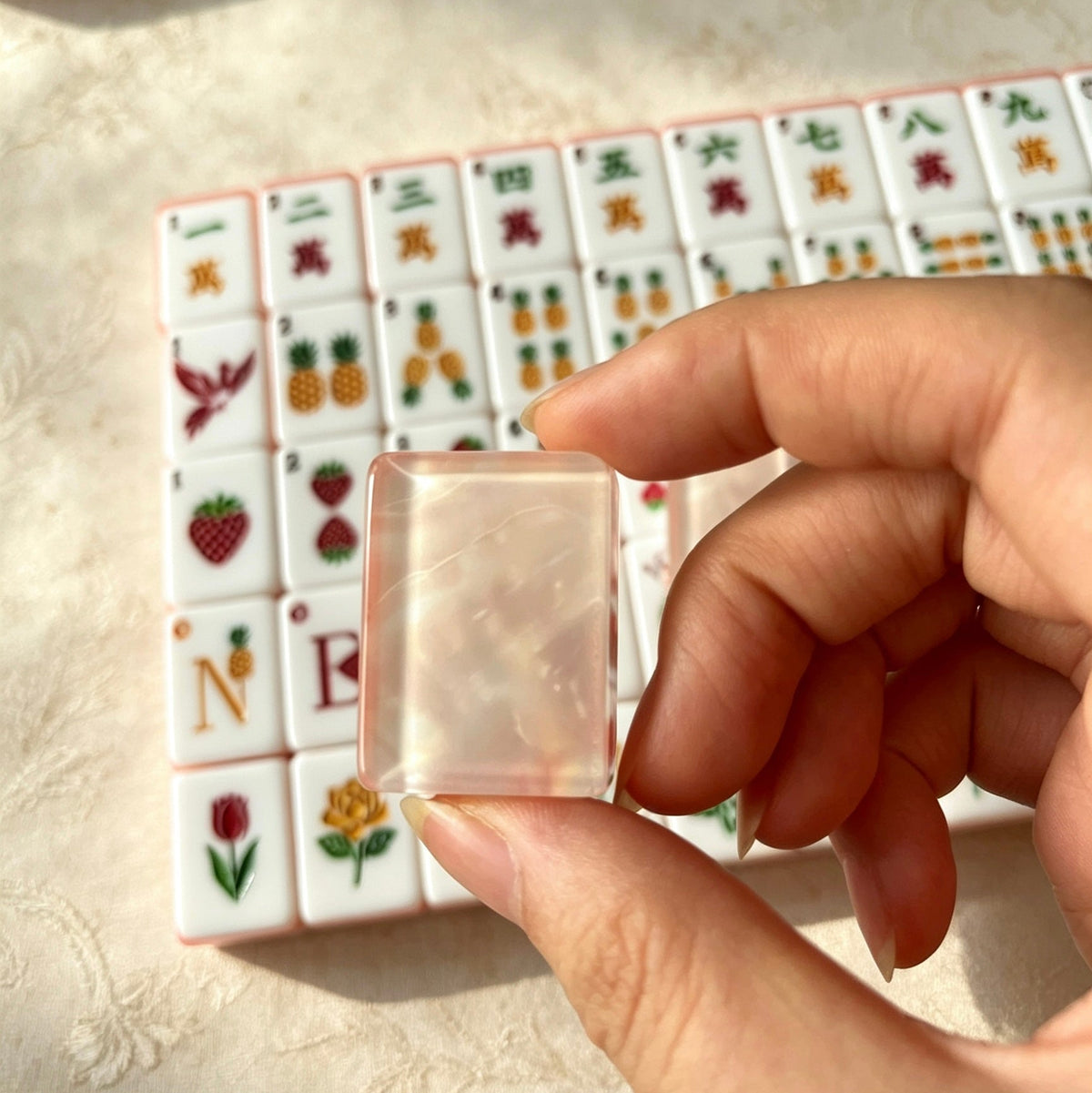 Scattered pile of Fruit Garden mahjong tiles, shimmering pink pearlescent backs, pastel pink-orange layered edges, strawberry and pineapple illustrated tile faces on linen surface