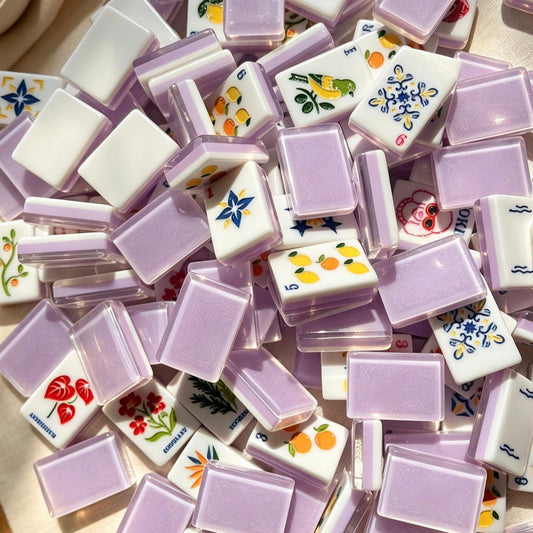 Lavender Fruit & Floral American mahjong tiles, translucent lavender purple resin backs, illustrated citrus fruit, floral and cartoon joker tiles, arranged on cream marble countertop with minimalist vases