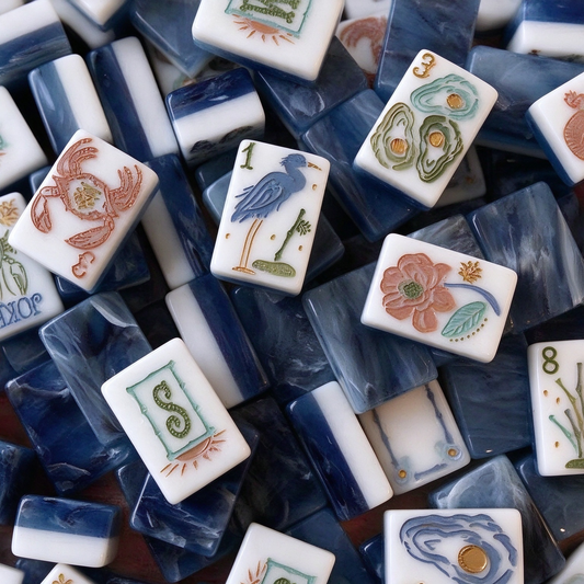 Immerse yourself in the wild beauty of southern coastal wetlands with this one-of-a-kind American mahjong set. Dreamy deep ocean-blue marbled tile backs pair with creamy white tile fronts, each hand-sculpted with intricate coastal wildlife artwork: crabs, alligators, herons, oysters and native marsh flora, reimagined to replace traditional mahjong symbols.