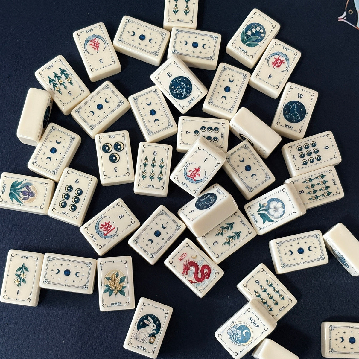 Side view of Moon Rabbit Tarot mahjong tiles lined up on acrylic rack over dark navy velvet fabric, cream ivory tiles with tarot border design, moon & star dots, floral bams, Chinese character cracks and moon rabbit lantern joker tile
