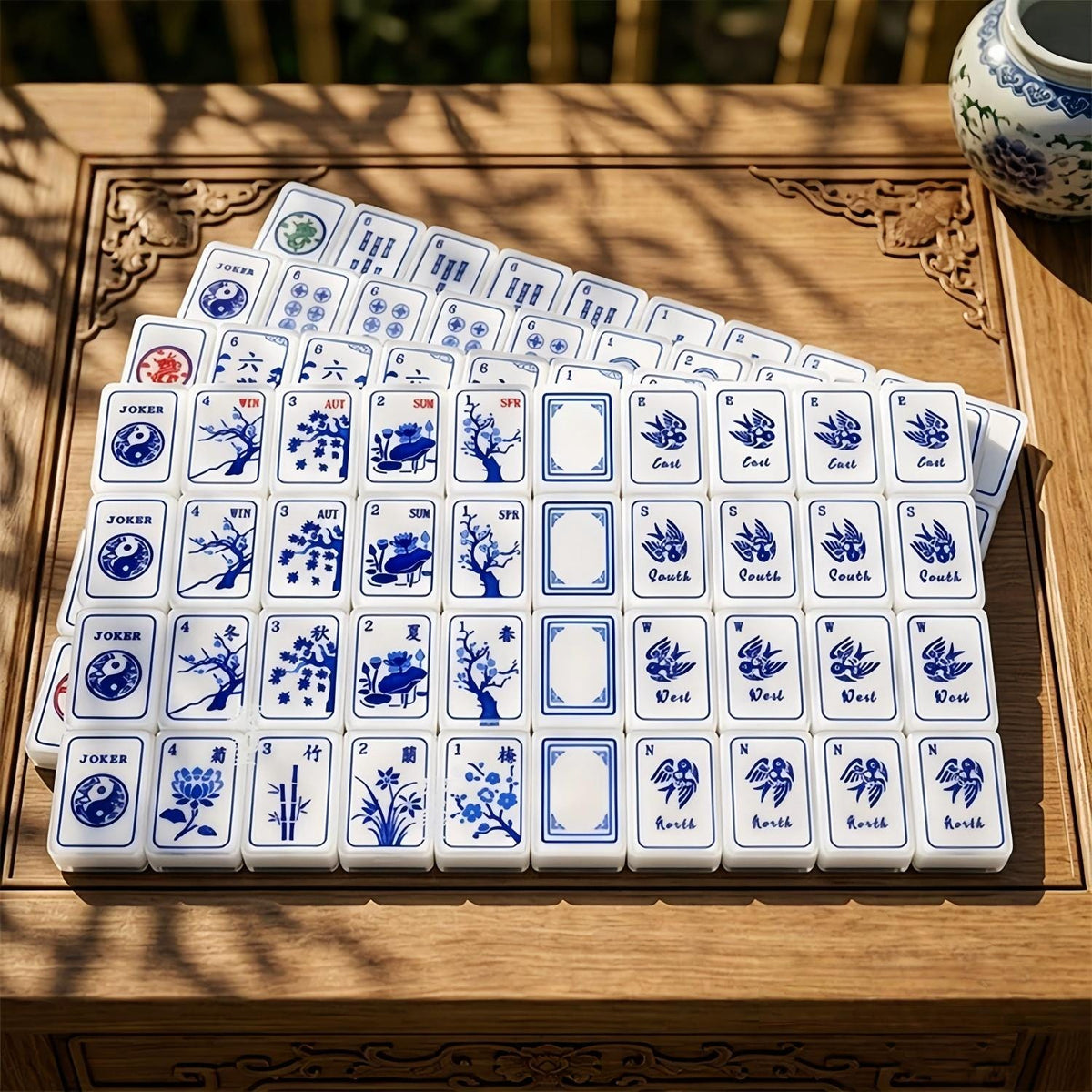Blue and white porcelain mahjong set displayed on wooden game table with matching blue porcelain mahjong mat, oriental garden background