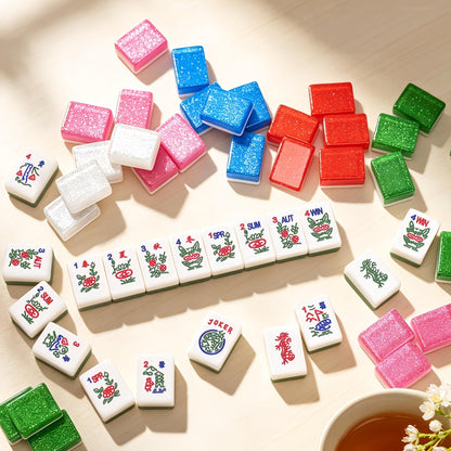 Crystal Shimmer mahjong set scattered on light wood table, translucent glitter tile backs in pink, blue, red, green and clear, white tile faces with season and flower artwork, colorful American mahjong tiles