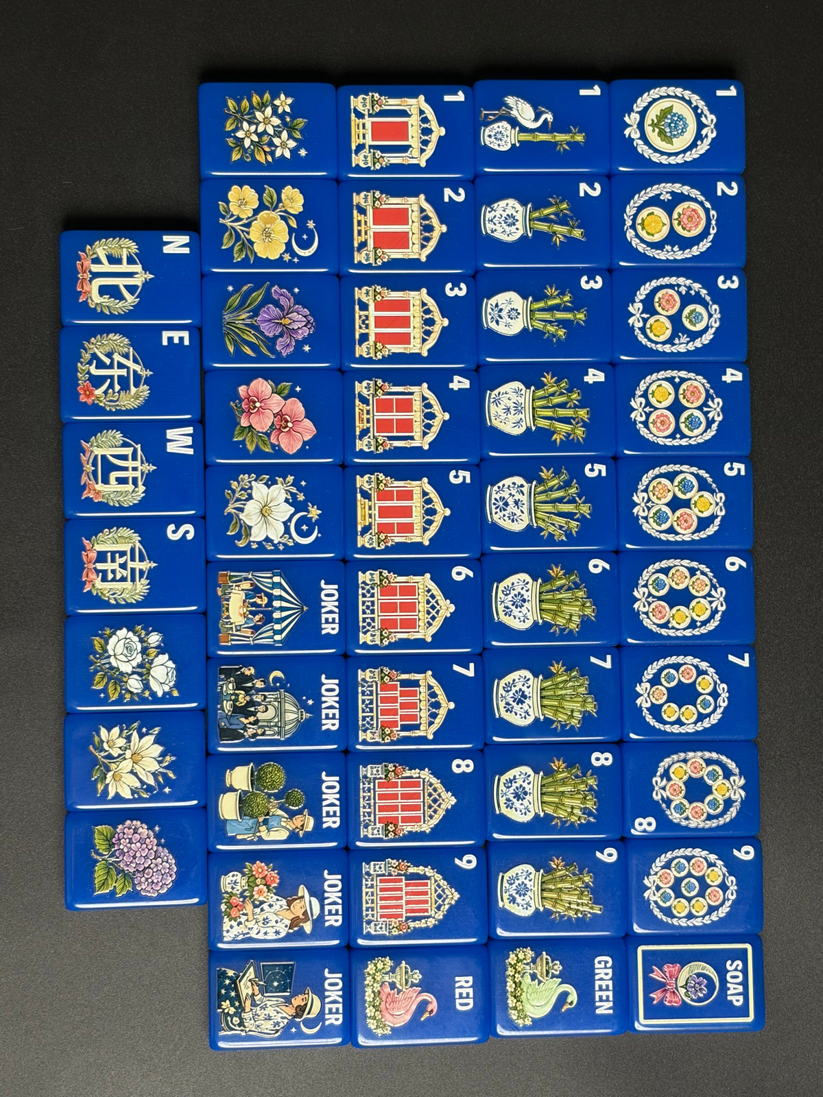 Top-down full layout of cobalt blue chinoiserie mahjong tiles arranged on black and white surface, showcasing porcelain vase bamboo, floral blooms, swan dragons and whimsical vintage joker artwork