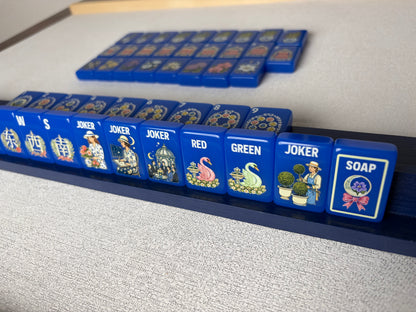 Side profile shot of Blue Chinoiserie mahjong tiles lined on dark wooden rack, glossy cobalt tiles displaying garden joker scenes, pink and white swan dragon tiles and floral SOAP tile